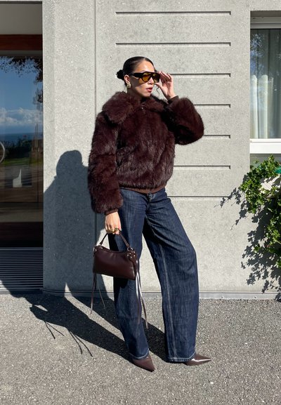 Mujer ajustándose grandes gafas de sol, vistiendo una chaqueta de piel marrón oscuro, jeans de pierna ancha, zapatos de punta y sosteniendo un pequeño bolso marrón en el exterior.