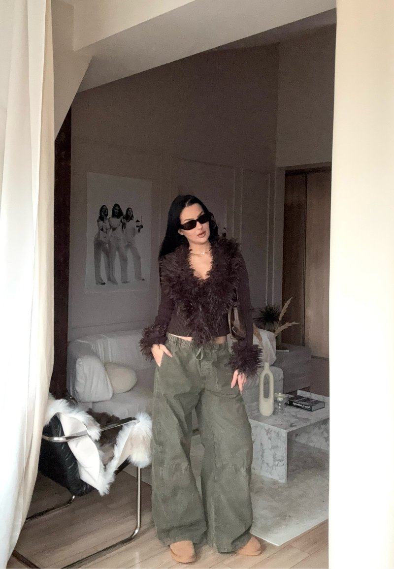 Braunes, gefiedertes Crop-Top mit langen Ärmeln, kombiniert mit lockeren olivgrünen Hosen. Accessoires sind schwarze Sonnenbrillen und eine braune Tasche.