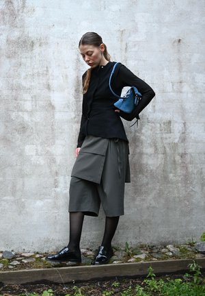 Schwarzes Langarmoberteil mit Knöpfen, graue weite Shorts mit Taschen, schwarze Socken und glänzende schwarze Schuhe. Kleine blaue Handtasche mit Riemen.