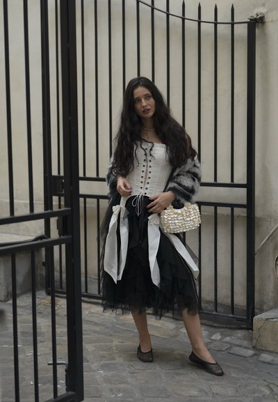Falda negra con volantes acompañada de una blusa blanca con cordones y un cárdigan de piel sintética gris. Un pequeño bolso de cuentas añade detalle. Unos zapatos planos de malla negra completan el look.