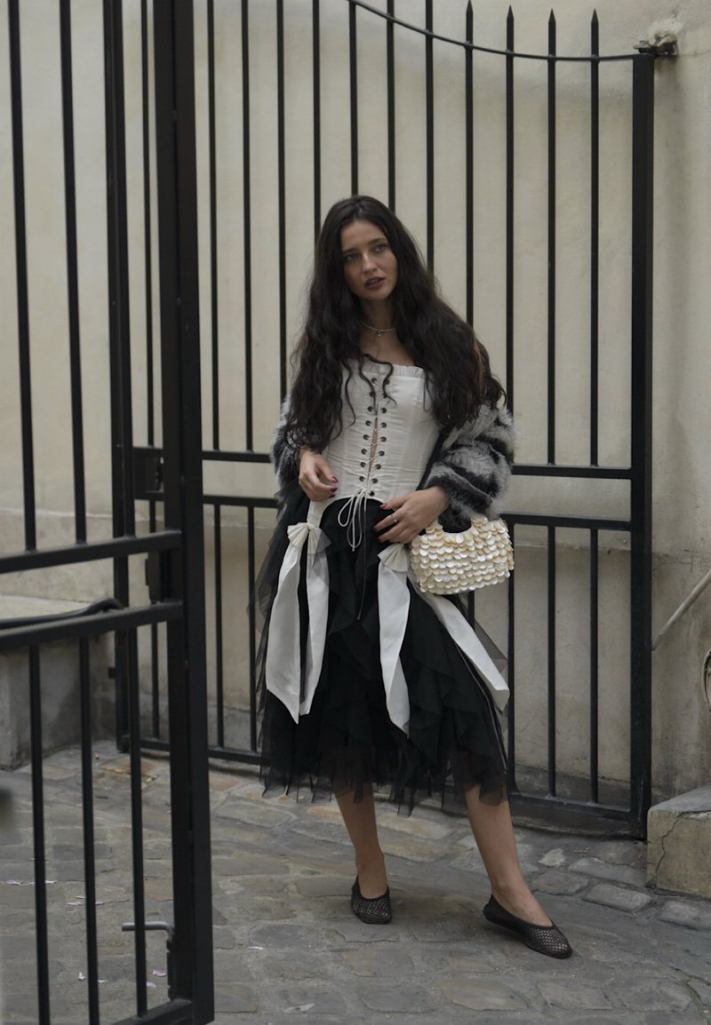 Crna volan suknja u kombinaciji s bijelim topom s čipkastim vezanjem i sivoj lažnoj krznoj cardigan. Mala torbica s perlicama dodaje detalj. Crne mrežaste balerinke upotpunjuju izgled.