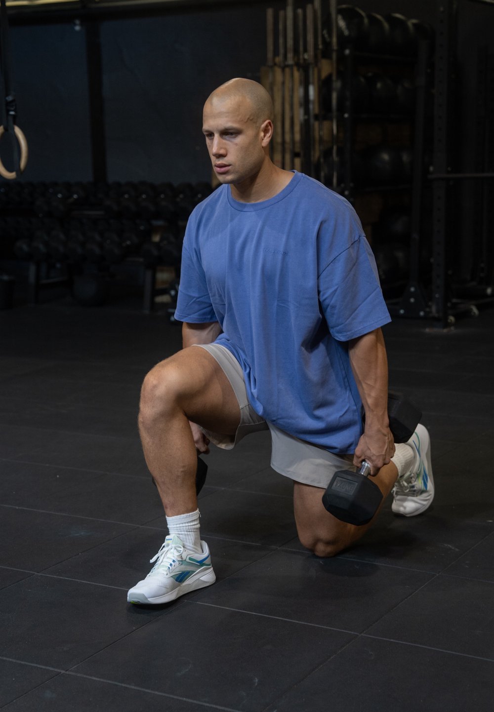 Un homme en T-shirt bleu surdimensionné et en shorts gris effectue un fente tout en tenant un haltère. Il porte des baskets blanches avec des accents bleus.