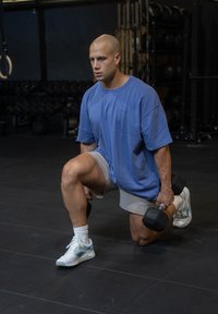 Un homme en T-shirt bleu surdimensionné et en shorts gris effectue un fente tout en tenant un haltère. Il porte des baskets blanches avec des accents bleus.