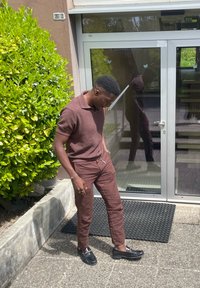 Camisa polo de punto marrón y pantalones a juego hechos de tejido texturizado, combinados con mocasines negros, de pie cerca de una puerta de cristal y vegetación.