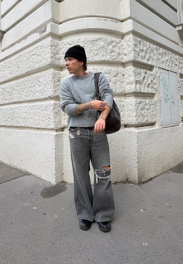 Joven con tatuajes que lleva un gorro negro, un suéter gris, vaqueros anchos rotos y zapatos negros, de pie junto a una pared de piedra con textura.