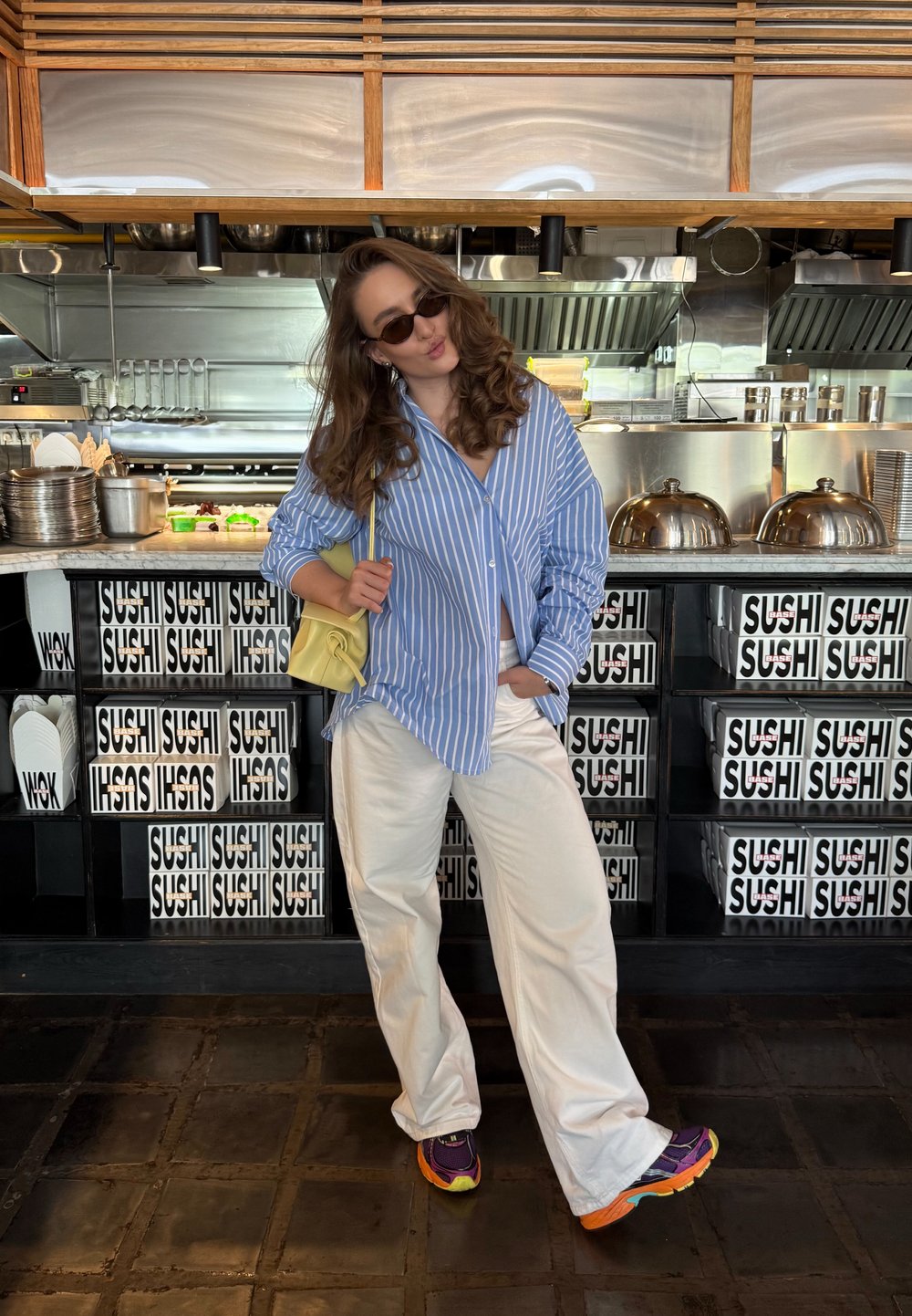 Blue and white striped button-up shirt, white wide-leg trousers, yellow bag, colourful sneakers; modern restaurant interior featuring sushi branding.