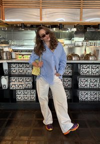 Blue and white striped button-up shirt, white wide-leg trousers, yellow bag, colourful sneakers; modern restaurant interior featuring sushi branding.