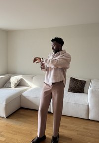 Homme ajustant sa montre, portant une veste et un pantalon rose pâle, debout dans un salon avec un canapé d'angle blanc et deux coussins, sol en bois.