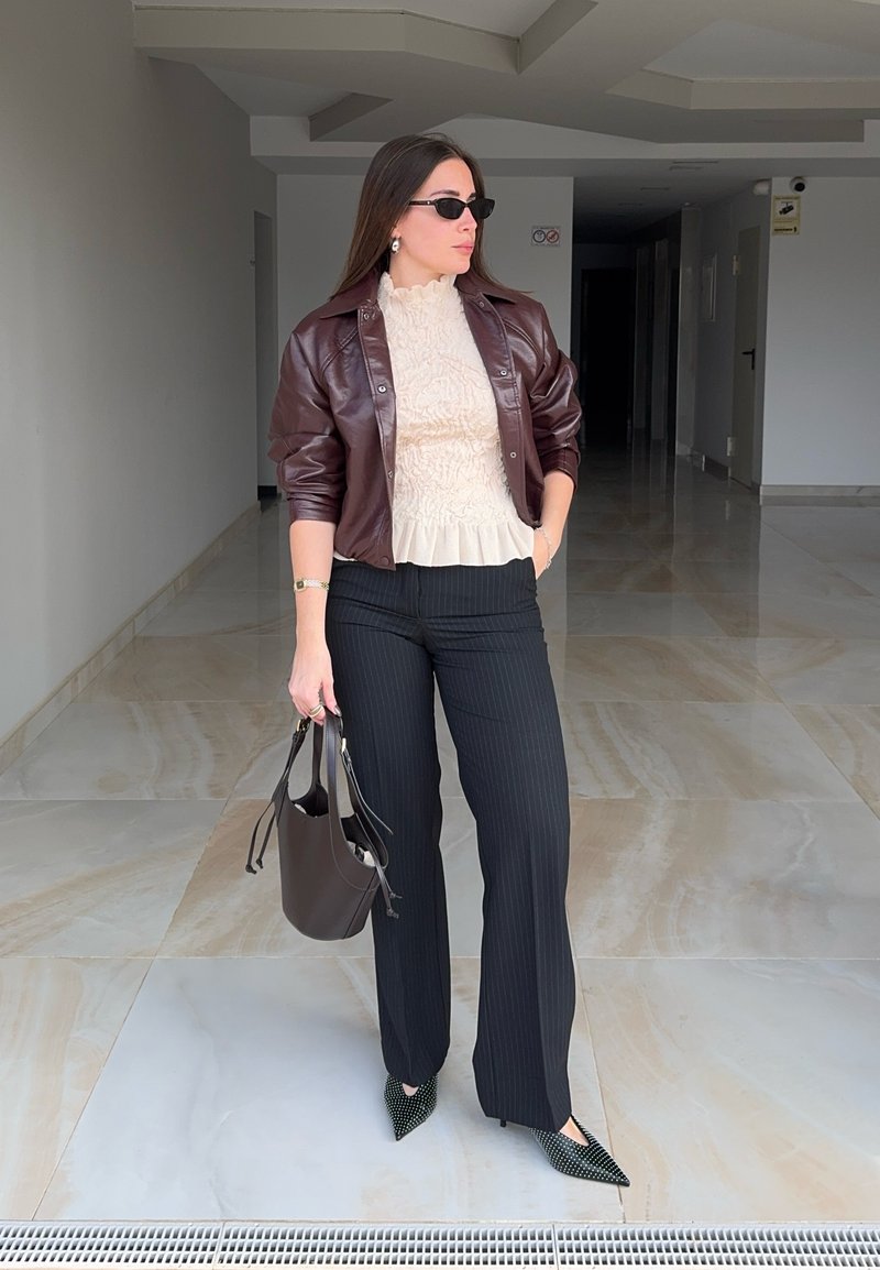 Femme portant des lunettes de soleil, veste en cuir marron, blouse crème, pantalon noir à fines rayures, tenant un sac à main sombre, debout dans un couloir carrelé.