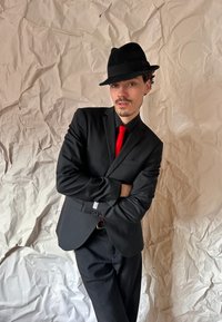 Man i svart kostym med röd slips och svart fedora, med armarna i kors, mot en strukturerad vit bakgrund.