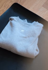 Sweatshirt cinza dobrado, feito de algodão, com um decote canelado e um pequeno logo branco da Adidas na parte da frente, colocado sobre uma superfície preta.