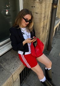 Schwarze Blazer, weißes T-Shirt mit einem Design, rote Sportshorts mit Streifen, schwarze kniehohe Stiefel und eine rote Tasche. Sonnenlicht beleuchtet die Szene.