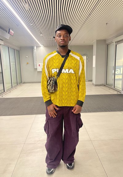 Camisa de manga larga amarilla con un patrón floral y el logo de "PUMA", combinada con pantalones cargo púrpuras holgados y zapatos negros. Incluye una pequeña mochila de camuflaje.