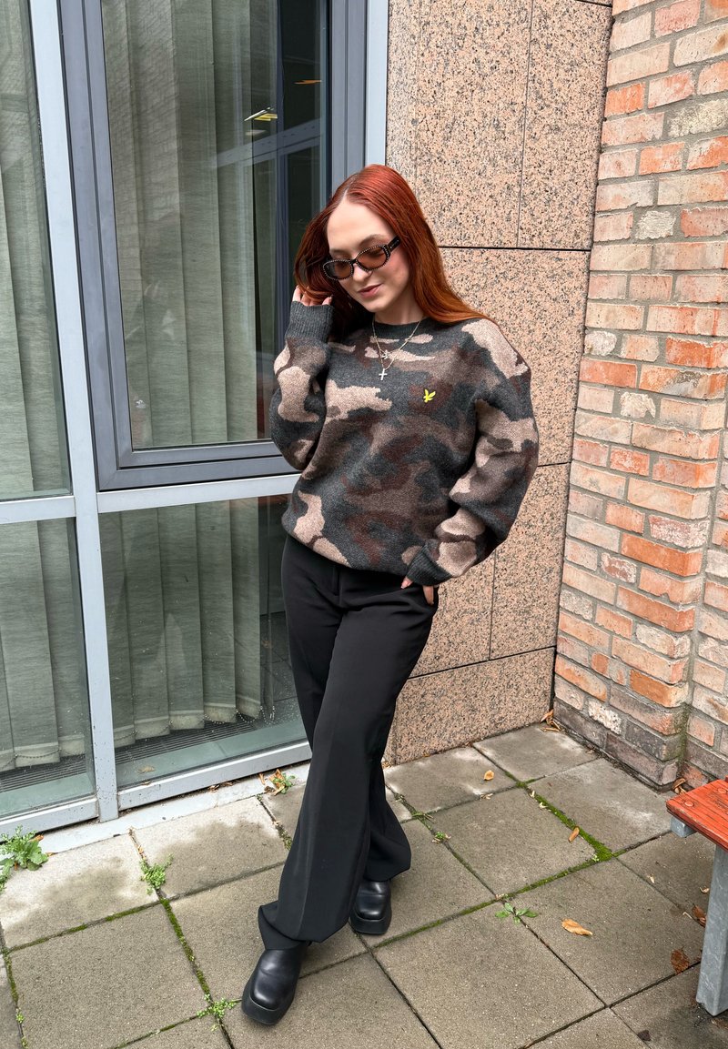 Pull en maille camouflage avec des nuances de marron et de gris, arborant un petit logo jaune. Pantalon noir à jambes larges et bottes noires épaisses.