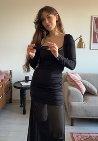 Melna pieguloša kleita ar gariem piedurknēm un caurspīdīgu apakšdaļu. Modelis tur melnas saulesbrilles. Fonā redzams dīvāns un dekoratīva lampa.