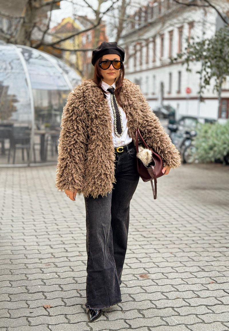 Femme portant un manteau marron duveteux, un béret noir, de grandes lunettes de soleil, une chemise blanche avec une cravate et un collier de perles, un pantalon noir, tenant un sac à main marron en extérieur.