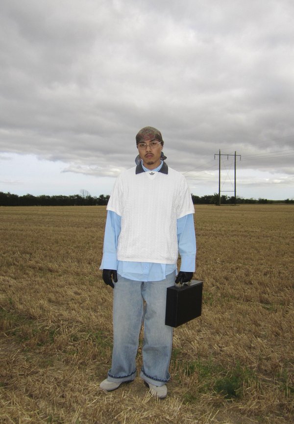 Mann mit Bandana, Brille, weißem Pullover und blauem Hemd steht mit einer schwarzen Aktentasche auf einem trockenen Feld unter bewölktem Himmel.