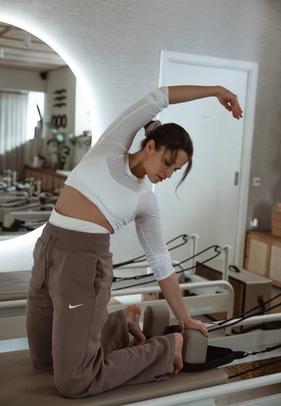 Reforma de pilates color crema, mujer con camiseta blanca de manga larga acanalada y pantalones de chándal topo, estirando el brazo por encima de la cabeza, descalza.