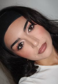 Portrait d'une personne ayant de longs cheveux foncés, portant un bandeau noir. La peau a un fini lisse ; le maquillage présente des teintes neutres avec des lèvres brillantes.