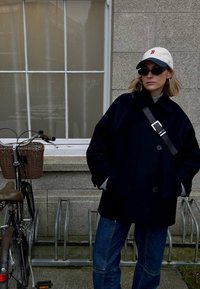 Femme portant des lunettes de soleil, une casquette blanche, un manteau sombre et un jean, debout les mains dans les poches à côté d'un vélo garé dans un support métallique contre un mur en pierre.
