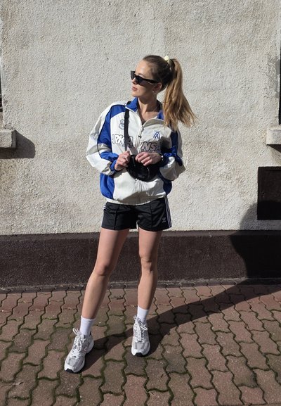 Rompevientos blanco y azul, pantalones cortos negros, zapatillas grises con acentos blancos. La modelo sostiene una cámara, estilizada con gafas de sol y una coleta.
