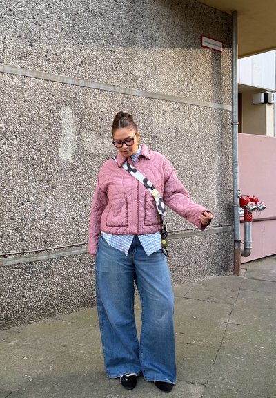 Mujer con chaqueta acolchada rosa, camisa azul a cuadros, vaqueros anchos, zapatos negros y gafas, de pie junto a una pared texturizada con hidrantes de incendios.