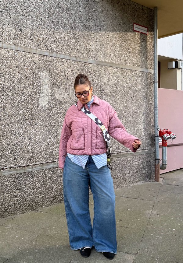 Femme portant une veste matelassée rose, une chemise à carreaux bleue, un jean large, des chaussures noires et des lunettes, se tenant près d'un mur texturé avec des bouches incendie.