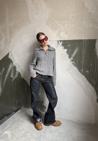 Grey ribbed knit jumper with a zip collar, black flared jeans, and tan boots. Oversized red sunglasses complete the outfit against a textured wall.