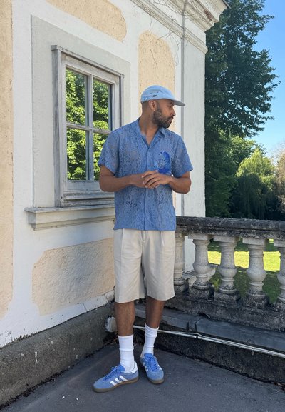 Camisa de encaje de manga corta color azul claro con un patrón texturizado, pantalones cortos crema, zapatillas azules y una gorra azul claro, de pie cerca de una barandilla de piedra.