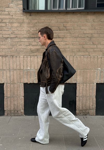 Hombre caminando por la acera junto a un muro de ladrillos, vestido con chaqueta de cuero marrón, pantalones de chándal grises, zapatos negros y llevando un gran bolso negro colgado al hombro.