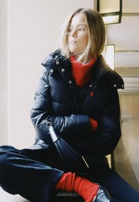 Ung kvinna med blont hår som bär en svart vadderad jacka, röd polotröja, röda strumpor och svarta skor, sittande med armarna i kors.