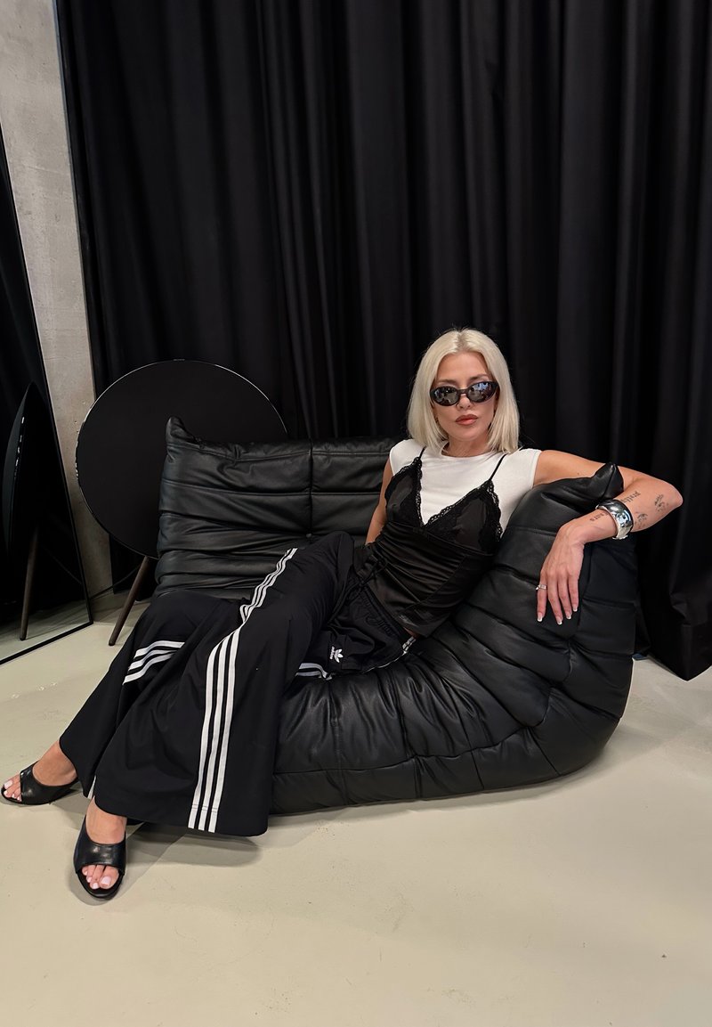 Femme aux cheveux blonds et lunettes de soleil se détendant sur un canapé en cuir noir, portant des vêtements noirs et blancs avec des bracelets argentés sur fond de rideau noir.