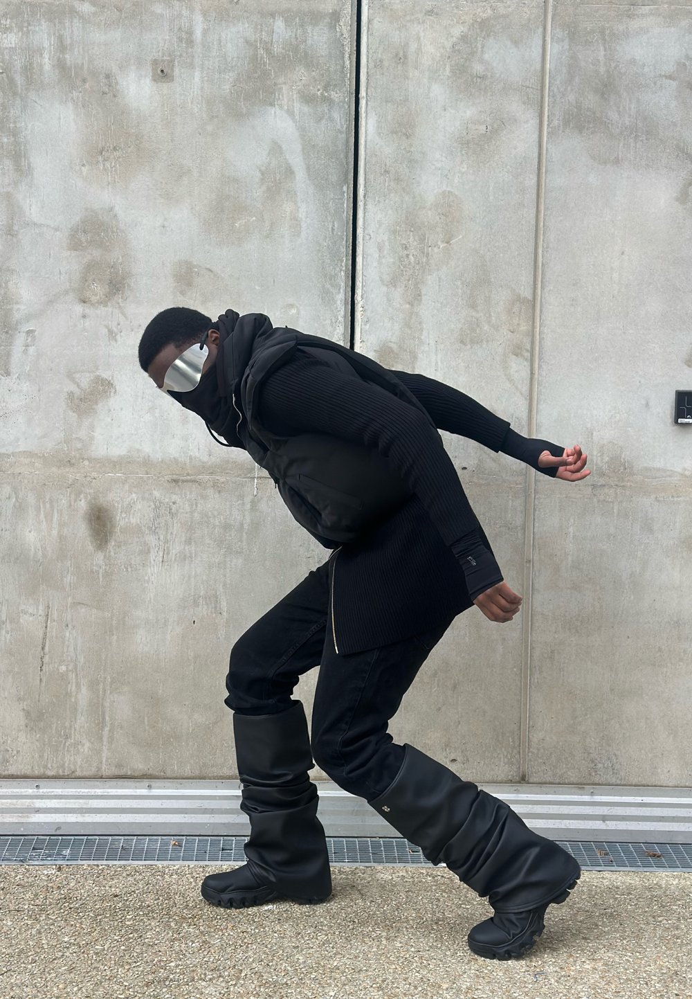 Person wearing a black puffer vest over a ribbed black sweater and relaxed-fit black jeans, paired with slouchy black boots and reflective sunglasses.