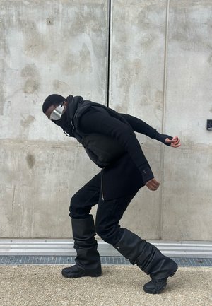 Persona vistiendo un chaleco acolchado negro sobre un suéter negro de canalé y jeans negros de corte relajado, combinado con botas negras holgadas y gafas de sol reflectantes.