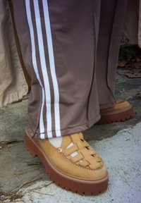 Pantaloni da ginnastica marroni con strisce bianche indossati sopra mocassini in pelle intrecciata beige, in piedi su una superficie di cemento crepato.