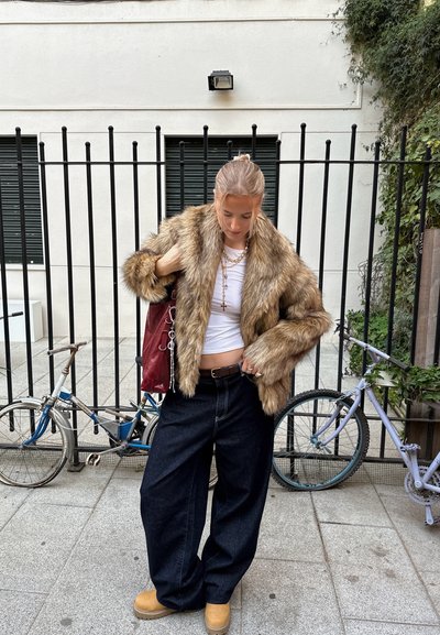 Abrigo de pelaje sintético en tonos marrones, pantalones de mezclilla negros de corte ancho, blusa blanca, joyería de cadena dorada y un bolso burdeos. Calzado con suela de madera.