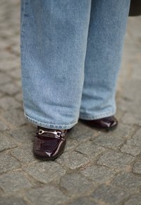 Burgunderfargede patent lærersko med spænde detaljer, parret med lyseblå vidde denim jeans. Gulvet dækket af grå brosten.