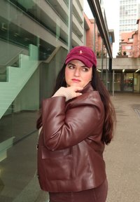 Braune Lederjacke mit weiter Passform und Fronttaschen, kombiniert mit einer burgunderroten Cordmütze und einem sichtbar beigen Pulloverärmeln.