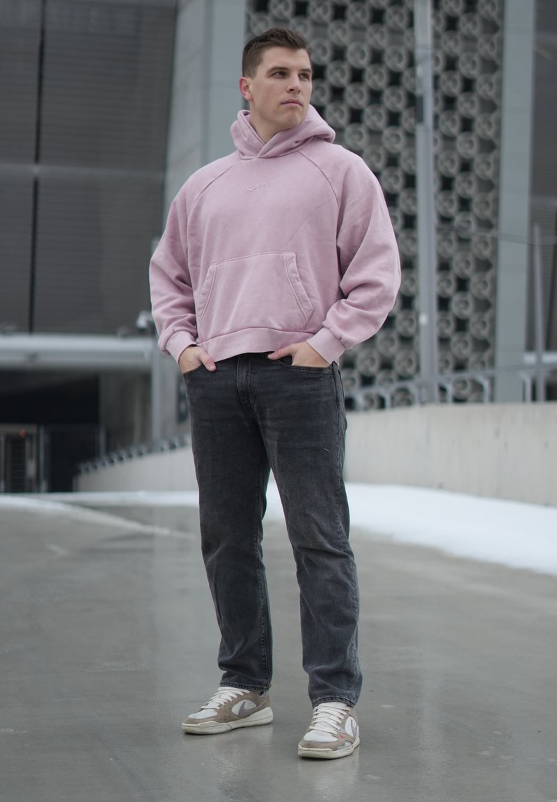 Jeune homme debout dans une rue urbaine portant un sweat à capuche rose, un jean noir et des baskets blanches, regardant sur le côté avec les mains dans les poches.