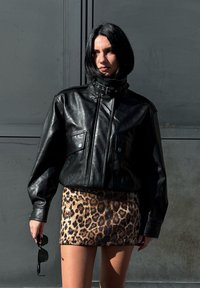 Crna kožna jakna s visokim ovratnikom i prednjim džepovima, uparena s uskom mini suknjom od leopard uzorka s šljokicama. Sunčane naočale drži u ruci.