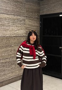 Pull rayé marron et blanc, accompagné d'une écharpe rouge, et d'une jupe marron foncé. La coupe est décontractée, avec une texture côtelée sur les manches.