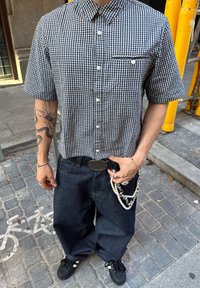 Kortermet checkered skjorte i marineblå og hvit, kombinert med mørke denimbukser. Tilbehøret inkluderer en kjede og armbånd.