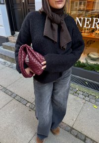 Čierny chlpatý sveter so širokými rukávmi, v kombinácii s voľnými sivými džínsovými nohavicami. Drží tkanú bordovú kabelku s zakriveným rukoväťou.