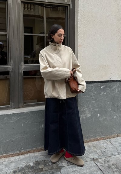 Chaqueta ligera beige, falda de mezclilla oscura con costuras laterales y bolso texturizado marrón. El atuendo incluye calzado gris y un fondo neutro.