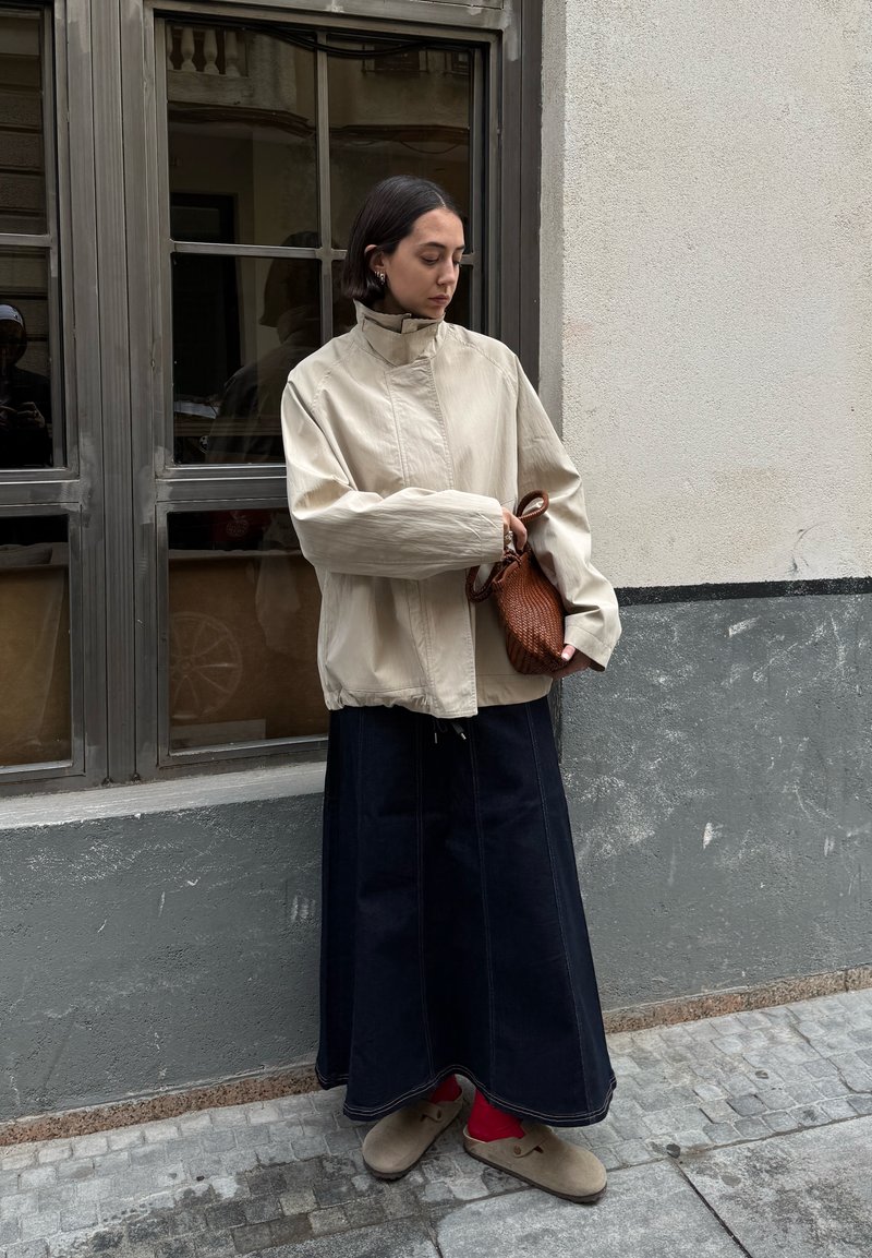 Beige leichte Jacke, dunkler Jeansrock mit Seitennähten und braune strukturiert Handtasche. Das Outfit beinhaltet graue Schuhe und einen neutralen Hintergrund.