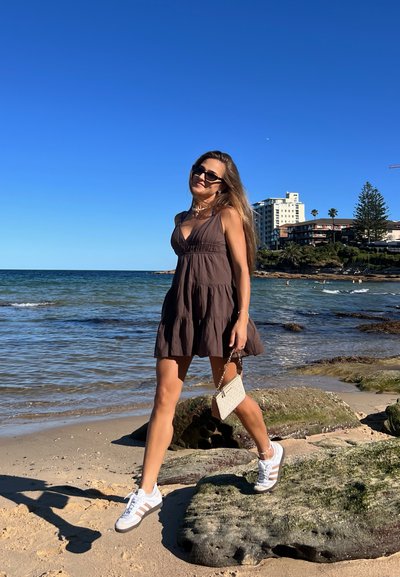 Vestido marrón sin mangas con falda acampanada, combinado con zapatillas blancas y un bolso pequeño. Playa arenosa y agua al fondo.