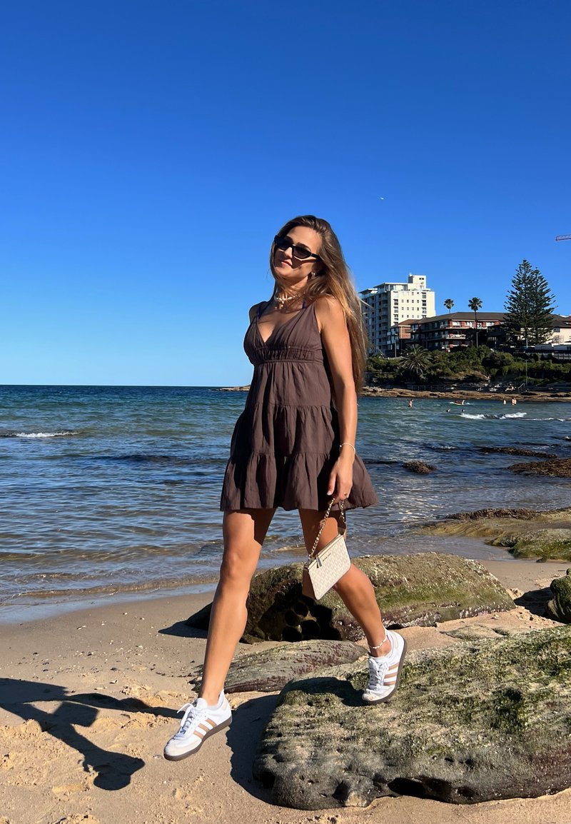 Braunes ärmelloses Kleid mit ausgestelltem Rock, kombiniert mit weißen Turnschuhen und einer kleinen Handtasche. Sandstrand und Wasser im Hintergrund.