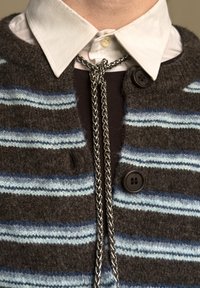 Chemise à col blanc sous un cardigan tricoté rayé marron et bleu, attaché par une chaîne argentée stylisée en cravate bolo.