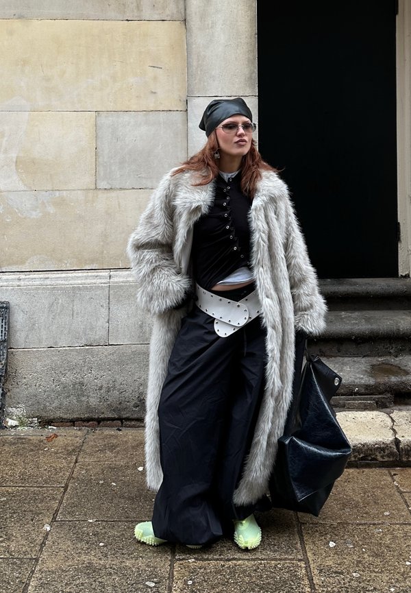 Femme portant un long manteau en fausse fourrure grise, haut noir, pantalon large, ceinture blanche à clous, foulard noir, lunettes de soleil, tenant un grand sac noir en extérieur.
