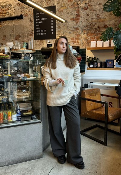 Suéter de gran tamaño color crema con dobladillo acanalado, combinado con pantalones de pierna ancha grises. Zapatos de plataforma negros, taza de cerámica minimalista y fondo de café rústico.